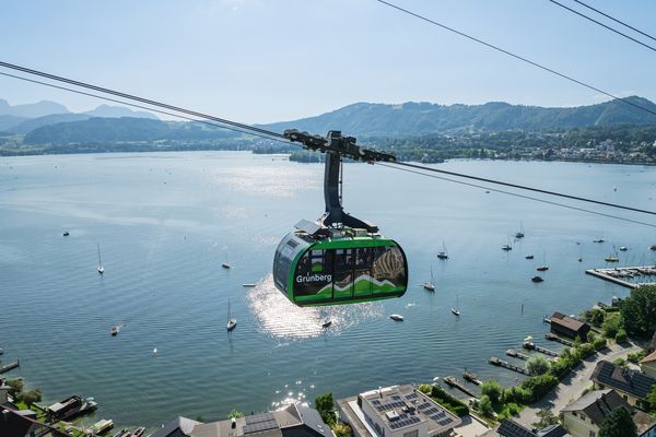 Grünberg-Bergbahn fährt über den Traunsee mit Segelbooten, grünen Hügeln und Blick auf die Stadt im Hintergrund
