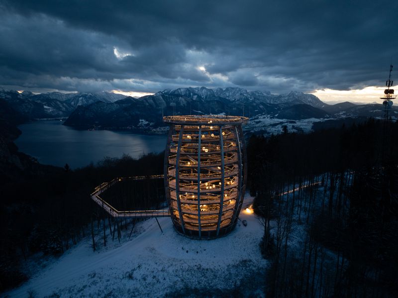 Spiraliger Turm mit Lichterketten in verschneitem Wald bei Nacht, Berge und See im Hintergrund.