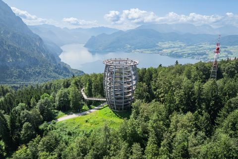 Spiralförmiger Holzaussichtsturm im dichten Wald mit Blick auf See und Berge unter blauem Himmel mit Wolken.