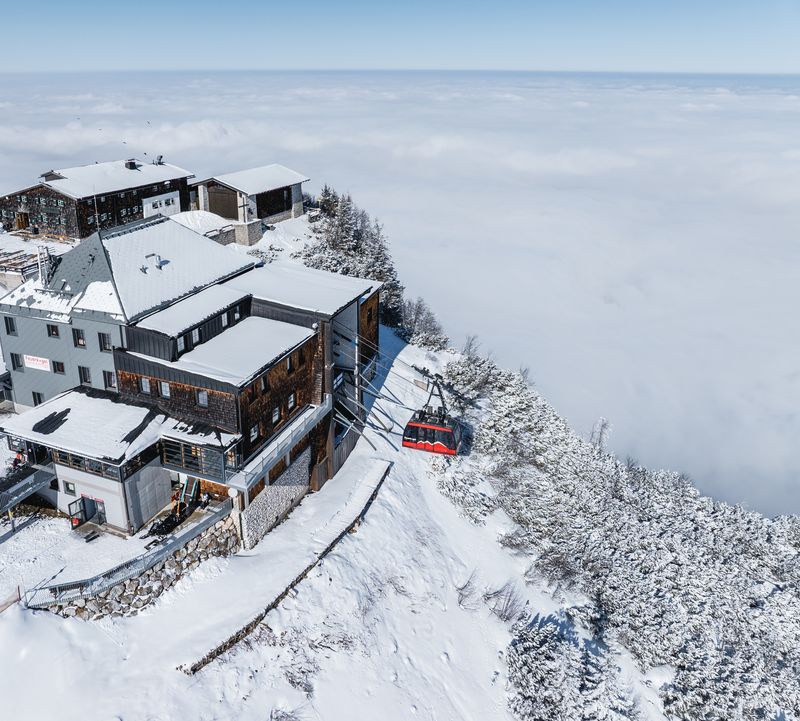 ie Feuerkogel-Bergstation liegt tief verschneit auf einem Plateau über dem Wolkenmeer, eine rote Seilbahn fährt gerade ein