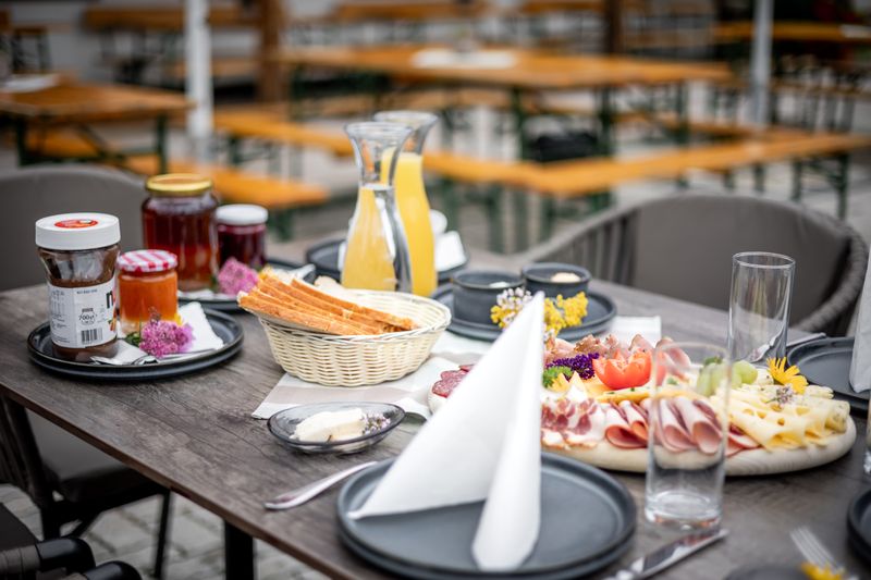 Ein Tisch im Freien mit einem Frühstücksbuffet, bestehend aus Aufschnitt, Käse, Brotstangen, Marmelade und einem Krug Orangensaft auf schwarzen Tellern.