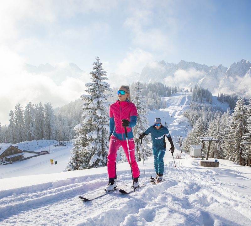 Zwei Personen gehen mit Skitourenski auf einen verschneiten Berg hinauf, umgeben von verschneiten Bäumen und entfernten Gipfeln unter einem klaren blauen Himmel.