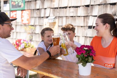 Familie sitzt an einem Holztisch im Sonnenschein und stößt mit Saft und Bier fröhlich an.