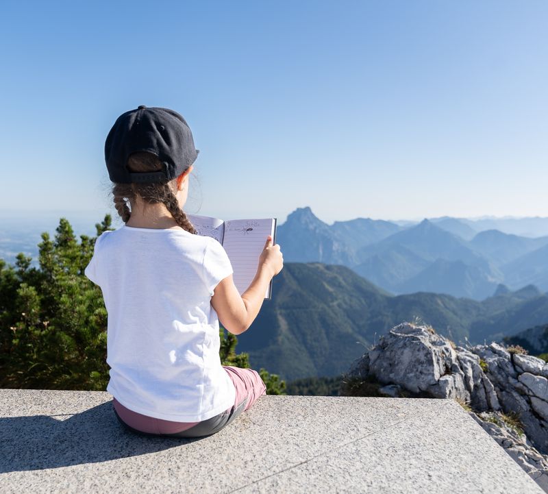 Mädchen sitzt mit offenem Gipfelbuch am Feuerkogel und blickt auf Traunstein und das weite Bergpanorama.