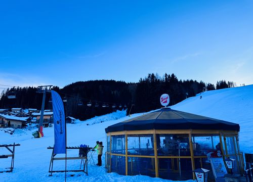 A snowy ski resort at dusk, featuring a circular building with glass walls, a chairlift, flags, and a forested hillside in the background.