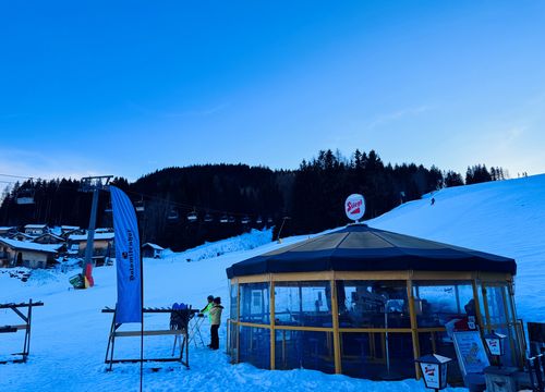 Ein verschneites Skigebiet in der Abenddämmerung mit einem runden Gebäude mit Glaswänden, einem Sessellift, Fahnen und einem bewaldeten Hügel im Hintergrund.
