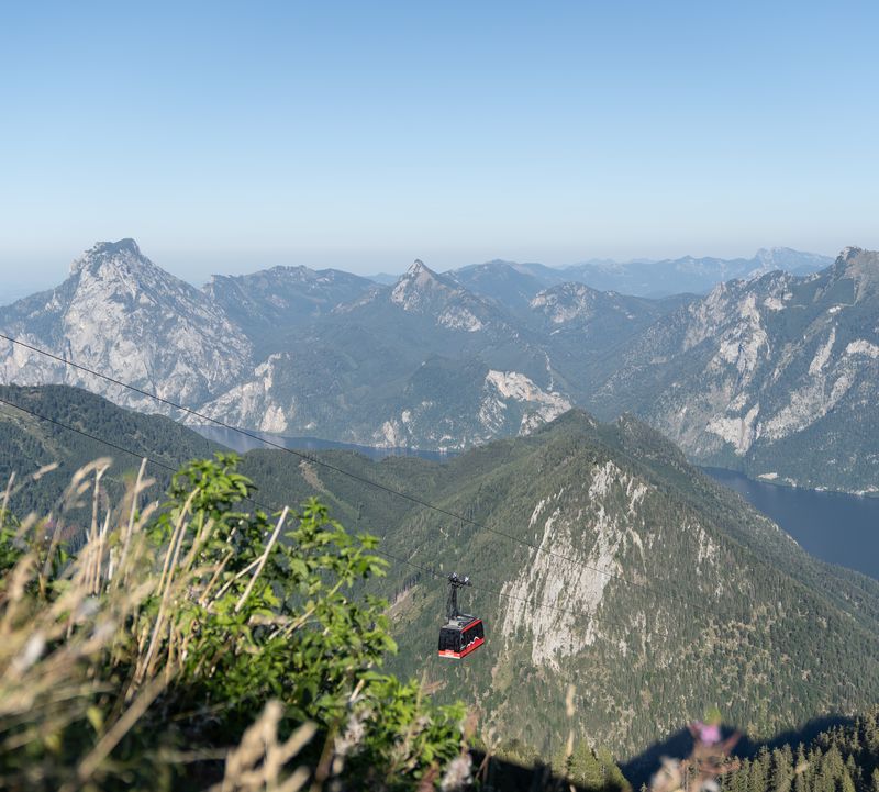 Die Bergbahn Feuerkogel schwebt über grüne Berghänge, mit Blick auf den Traunsee und schroffe Gipfel, hinauf auf den Feuerkogel.