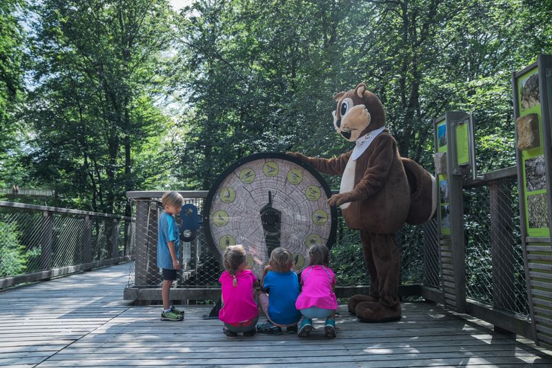 Kinder spielen mit einem Eichhörnchen-Maskottchen neben einer großen Uhr auf einem Holzweg im Wald.