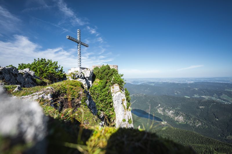 Das Europakreuz steht auf einem Felsvorsprung mit Blick ins Alpenvorland, der Himmel ist strahlend blau.
