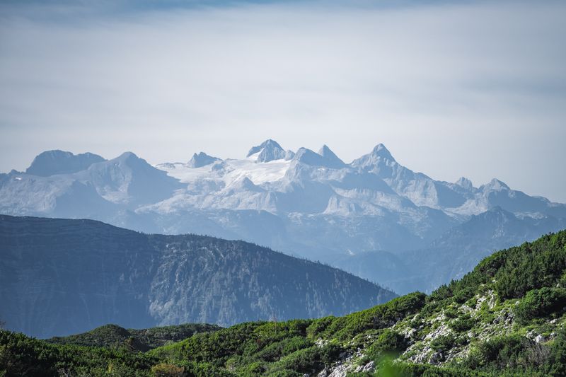 Schneeüberzogene Gipfel des Dachsteinmassivs ragen über grüne Berghänge und dunkle Wälder.
