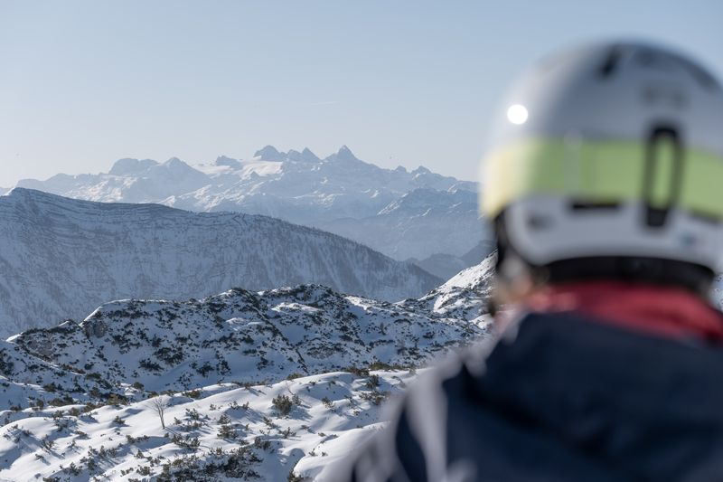 Eine Person mit Skihelm blickt über verschneite Hänge auf das markante Dachsteinmassiv in der Ferne.