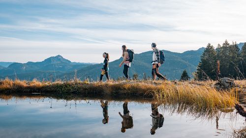 Drei Wanderer gehen auf einem Pfad neben einem spiegelnden See, umgeben von goldbraunem Gras und einer Berglandschaft unter blauem Himmel.
