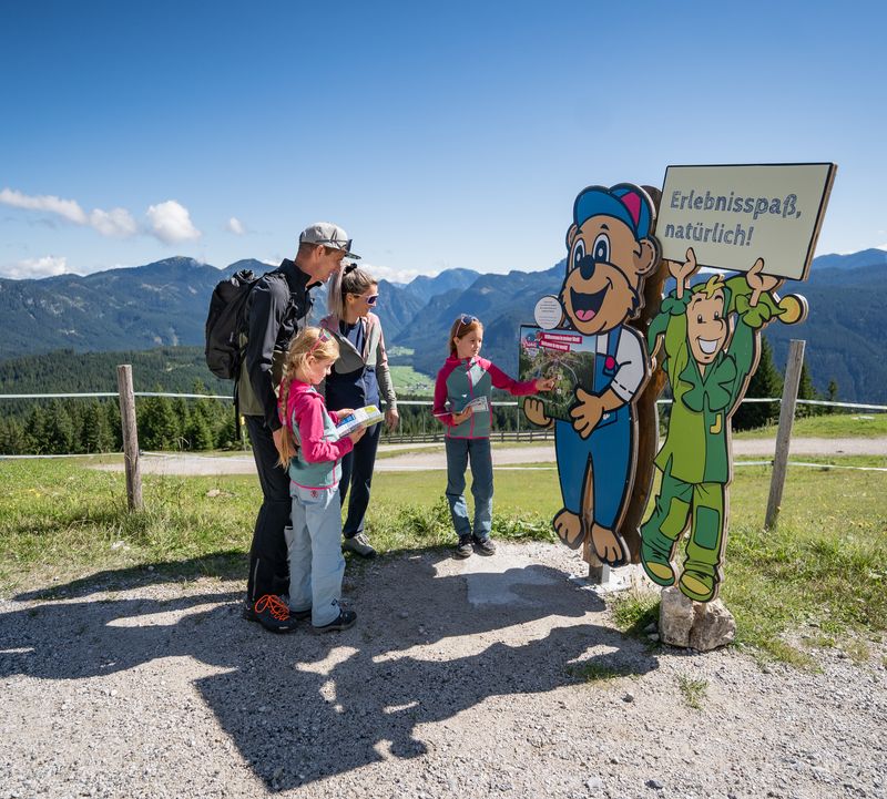 Familie auf einem Bergpfad, die bei blauem Himmel vor zwei bunten Cartoon-Figuren mit Schildern stehen. Die grüne Jolly-Figur hält ein Schild mit der Aufschrift „Erlebnisspaß natürlich!“ in die Höhe. Ein Kind drückt auf dem Schild bei der Bären-Figur auf einen Knopf.