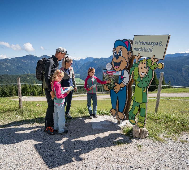 A family on a mountain path standing in front of two colourful cartoon characters holding signs against a blue sky. The green Jolly character is holding up a sign that reads ‘Fun and adventure, naturally!’ A child is pressing a button on the sign next to the bear character.