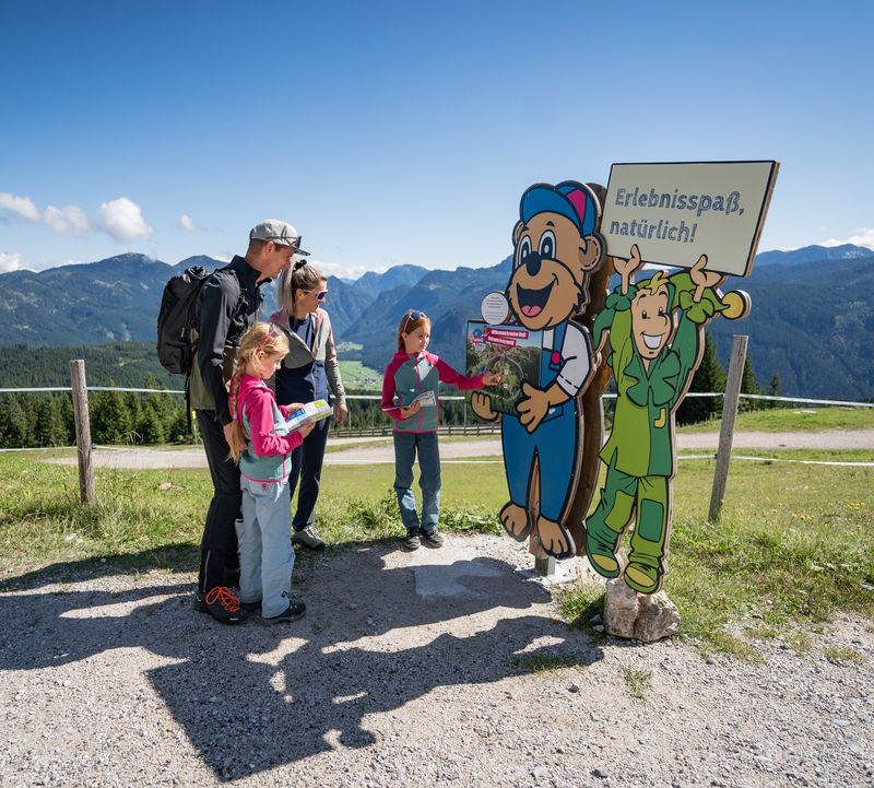 Familie auf einem Bergpfad, die bei blauem Himmel vor zwei bunten Cartoon-Figuren mit Schildern stehen. Die grüne Jolly-Figur hält ein Schild mit der Aufschrift „Erlebnisspaß natürlich!“ in die Höhe. Ein Kind drückt auf dem Schild bei der Bären-Figur auf einen Knopf.