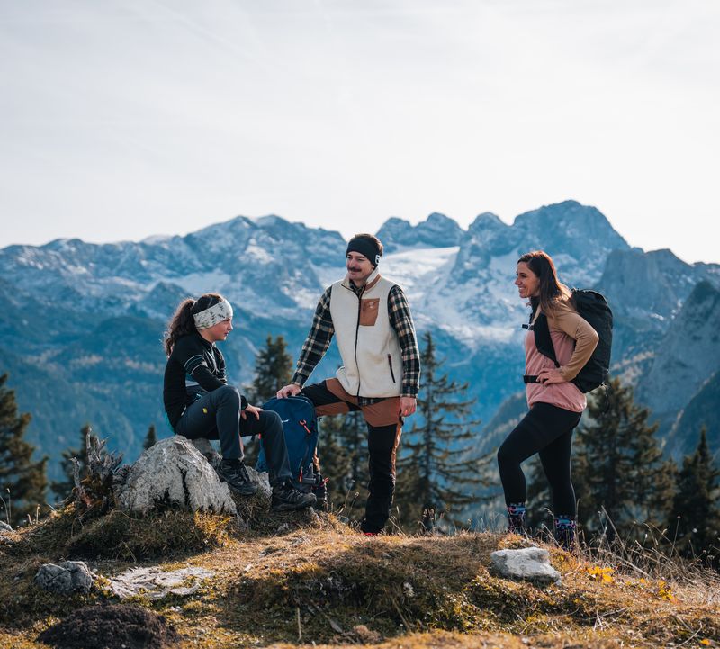 Drei Wanderer machen Pause auf einem Hügel mit Blick auf schneebedeckte Alpen und Kiefern unter blauem Himmel.