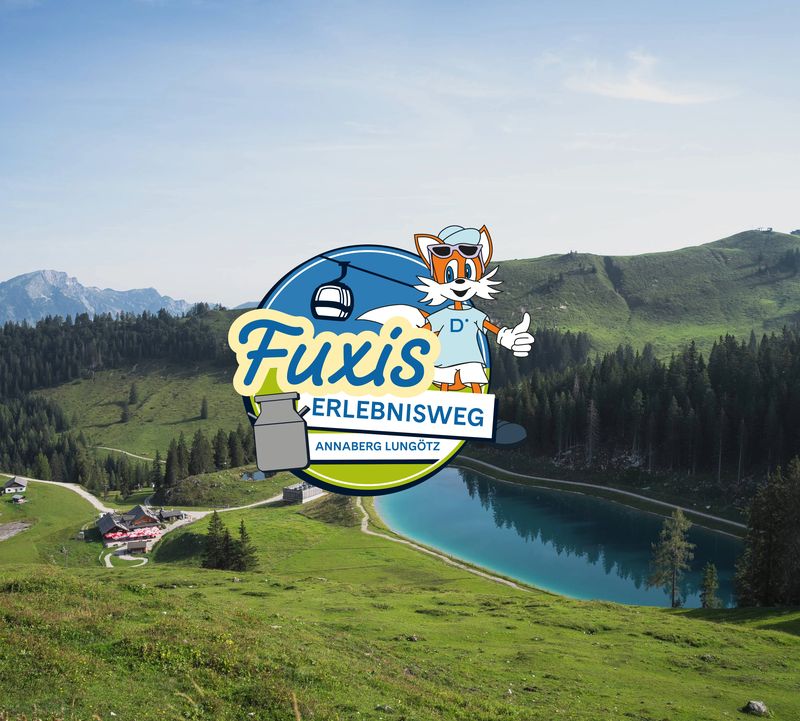 Das Logo des „Fuxis Erlebnisweg“ mit einem Comic-Fuchs, einem Skilift und einem Milchkännchen, eingebettet in eine malerische Berglandschaft mit einem See und grünen Hügeln.