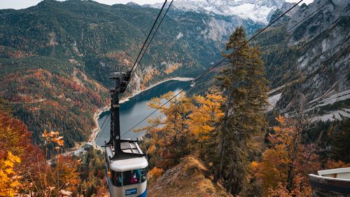 Eine Seilbahnkabine schwebt über einem tiefblauen See, umgeben von herbstlichen Wäldern und schneebedeckten Bergen.
