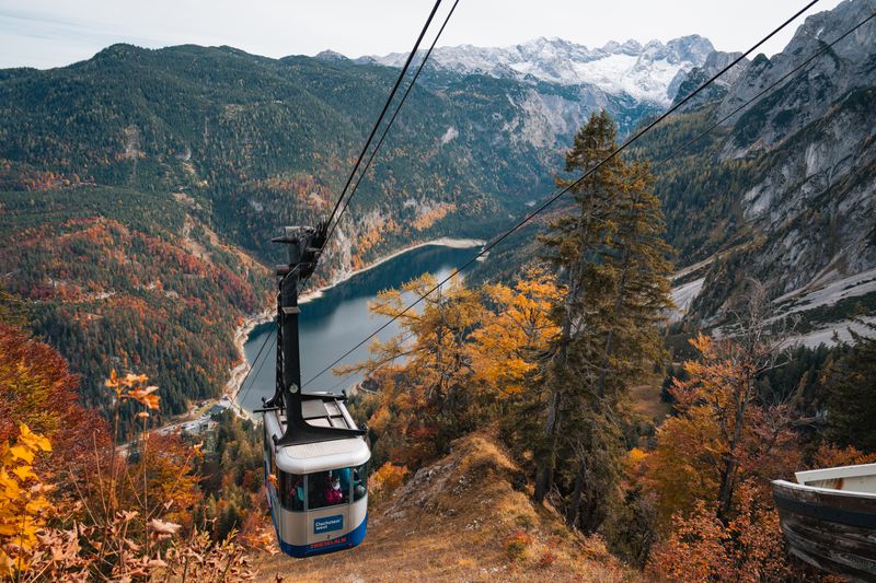 Eine Seilbahnkabine schwebt über einem tiefblauen See, umgeben von herbstlichen Wäldern und schneebedeckten Bergen.