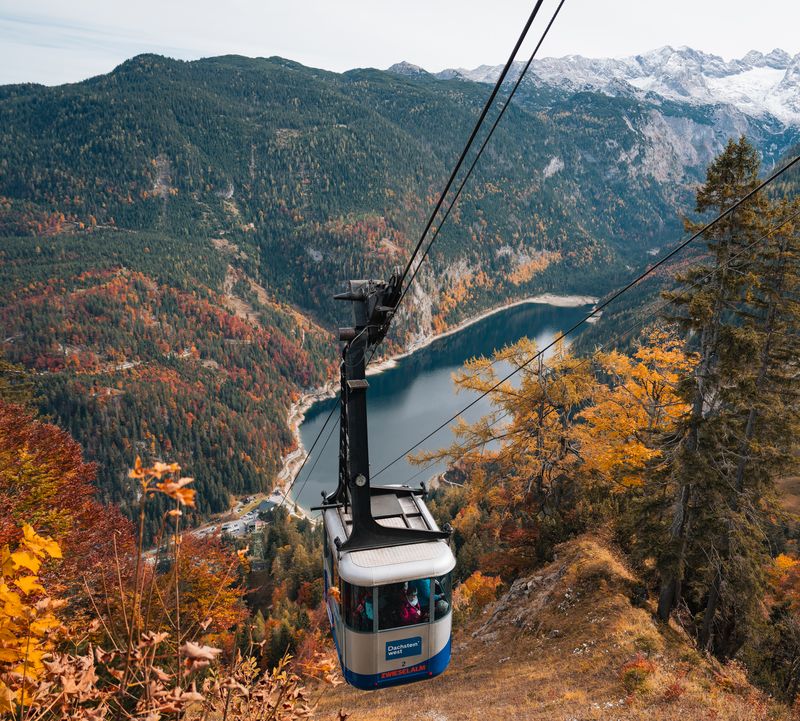 A cable car cabin floats above a deep blue lake, surrounded by autumnal forests and snow-covered mountains.