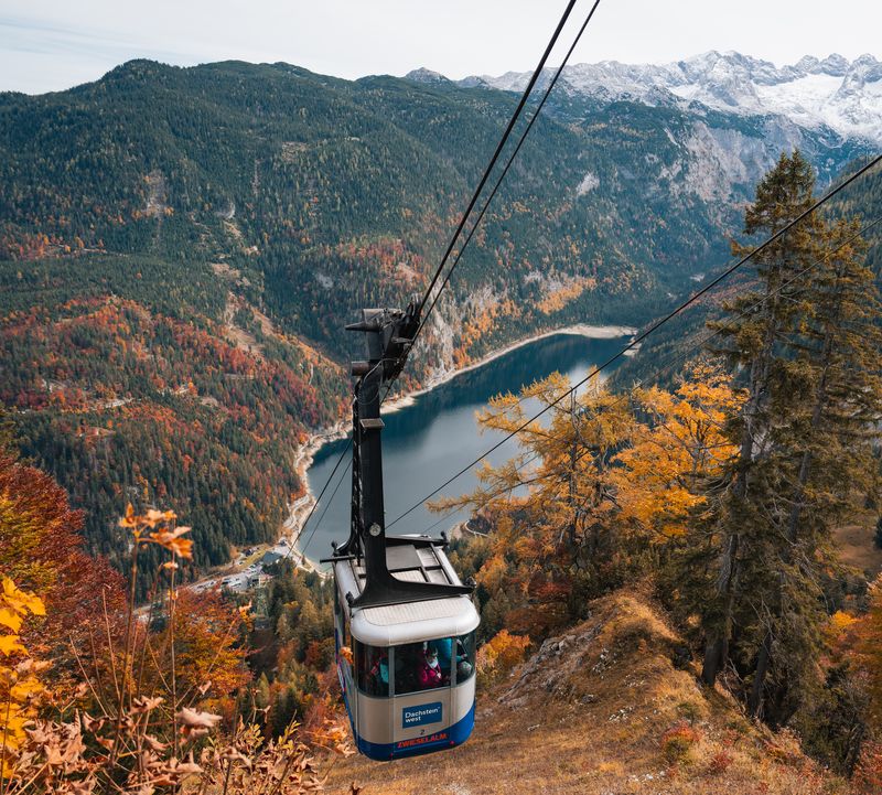 Eine Seilbahnkabine schwebt über einem tiefblauen See, umgeben von herbstlichen Wäldern und schneebedeckten Bergen.