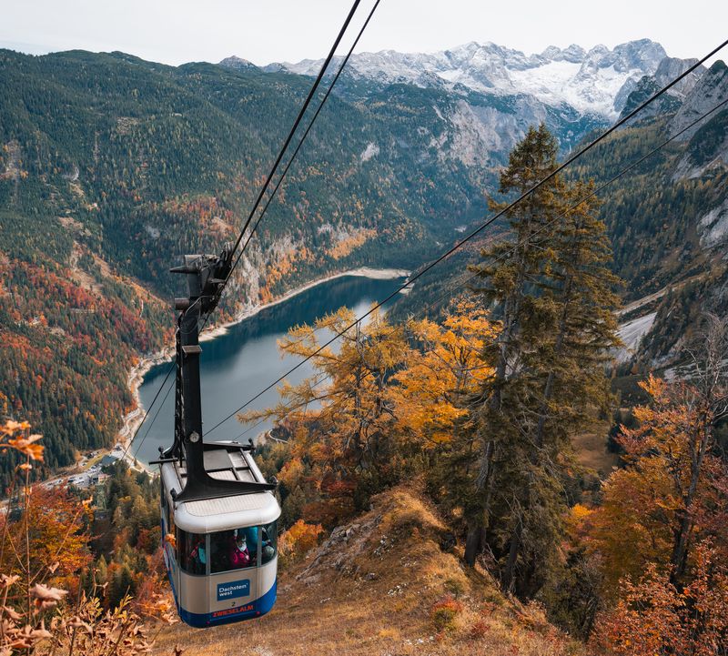 Eine Seilbahnkabine schwebt über einem tiefblauen See, umgeben von herbstlichen Wäldern und schneebedeckten Bergen.