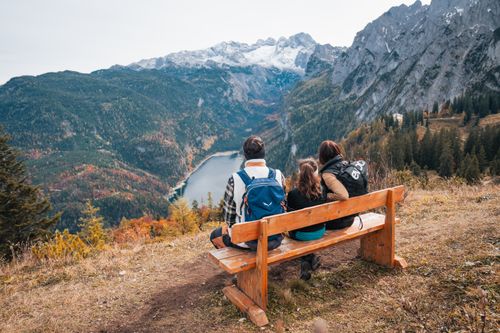 A family on a bench enjoys the sweeping view across a mountain lake and autumnal forests to snow-capped peaks.