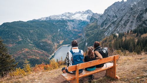 Familie auf einer Bank genießt den weiten Blick über einen Bergsee und herbstliche Wälder zu schneebedeckten Gipfeln.