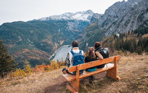 Familie auf einer Bank genießt den weiten Blick über einen Bergsee und herbstliche Wälder zu schneebedeckten Gipfeln.
