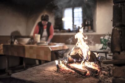 Loderndes Holzfeuer auf einem Steinofen im Vordergrund, während im unscharfen Hintergrund eine Person in einer rustikalen Küche arbeitet.