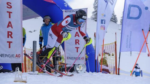 Ein Snowboarder in farbenfrohen Rennanzug macht eine scharfe Kurve um eine blaue Riesentorfahne auf einer Skipiste unter einem klaren blauen Himmel.