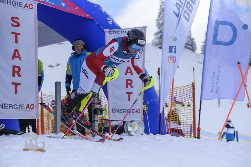Ein Snowboarder in farbenfrohen Rennanzug macht eine scharfe Kurve um eine blaue Riesentorfahne auf einer Skipiste unter einem klaren blauen Himmel.