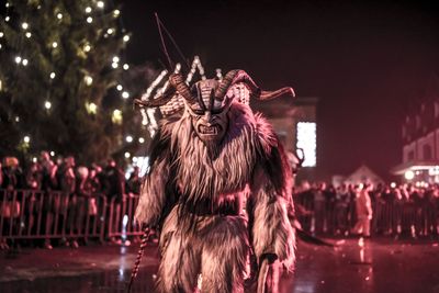 Furchterregender Krampus mit Hörnern und zotteligem Fell steht nachts vor Zuschauern und leuchtenden Festlichtern.