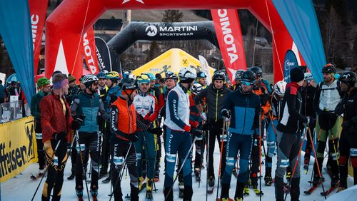 Eine Gruppe von Skitourengeher in bunter Ausrüstung steht an einer verschneiten Startlinie unter einem roten aufblasbaren Bogen und bereitet sich auf ein Rennen vor.