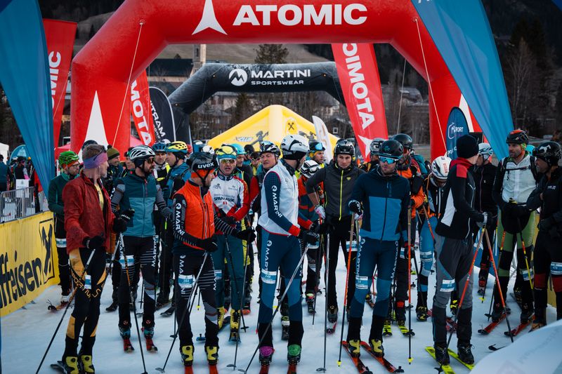 Eine Gruppe von Skitourengeher in bunter Ausrüstung steht an einer verschneiten Startlinie unter einem roten aufblasbaren Bogen und bereitet sich auf ein Rennen vor.