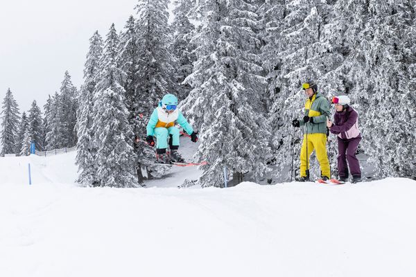A child in light blue ski clothing performs a jump in the air while two people watch from a snowy slope surrounded by snow-covered trees.