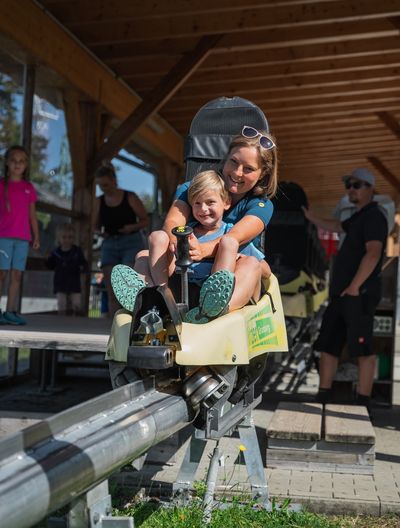 Eine Frau und ein Kind lachen, während sie in einer der Grünberg Sommerrodeln starten, mit Zuschauern im Hintergrund.