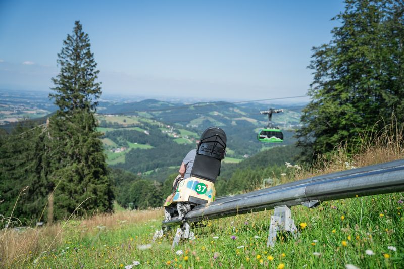 Eine Person fährt mit der Sommerrodelbahn am Grünberg, mit Grünberg Seilbahn und Berglandschaft im Hintergrund.