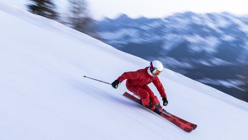 A skier in a red outfit and helmet swiftly descends a snowy slope with blurred mountains in the background.