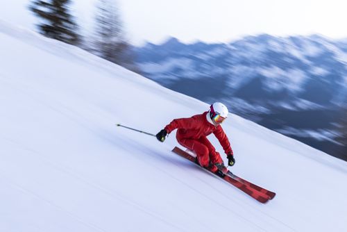 A skier in a red outfit and helmet swiftly descends a snowy slope with blurred mountains in the background.