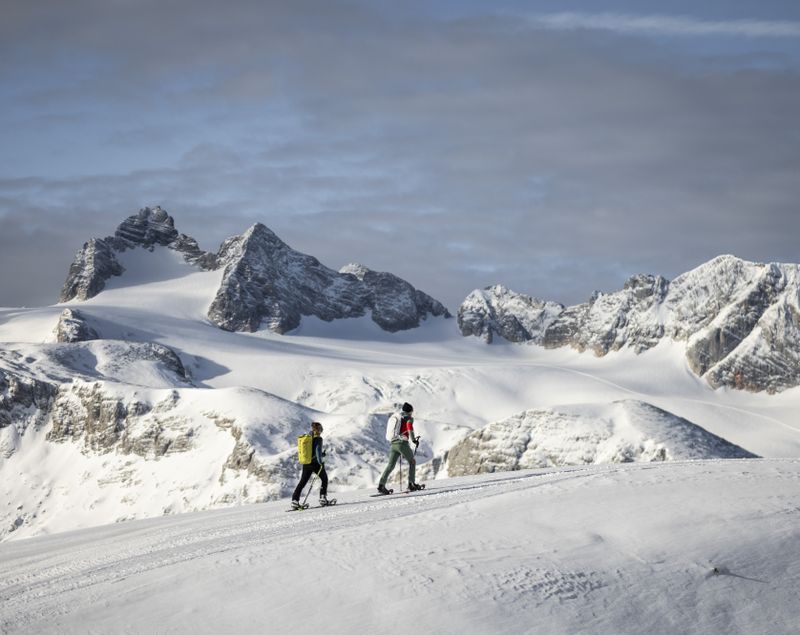 Zwei Schneeschuhwandernde gehen durch tiefen Schnee mit Blick auf das Dachsteinmassiv.