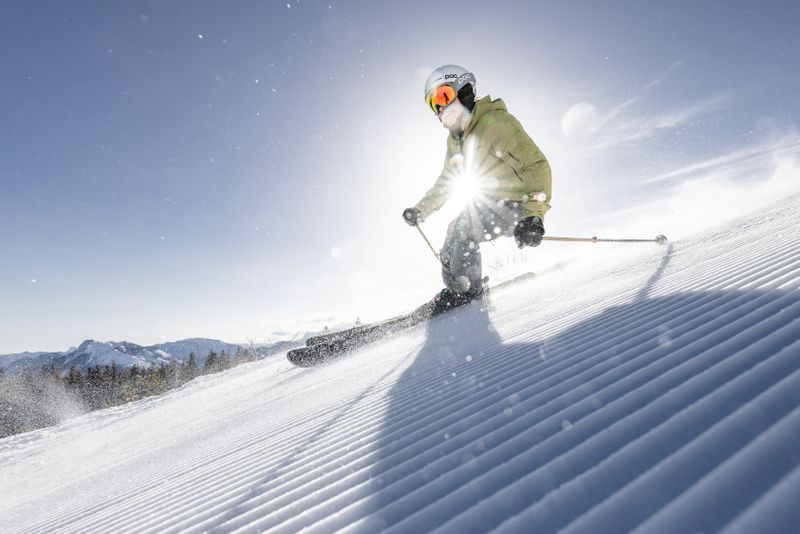 Skier in a green jacket carves dynamically down a freshly groomed slope at sunrise, snow crystals flying.