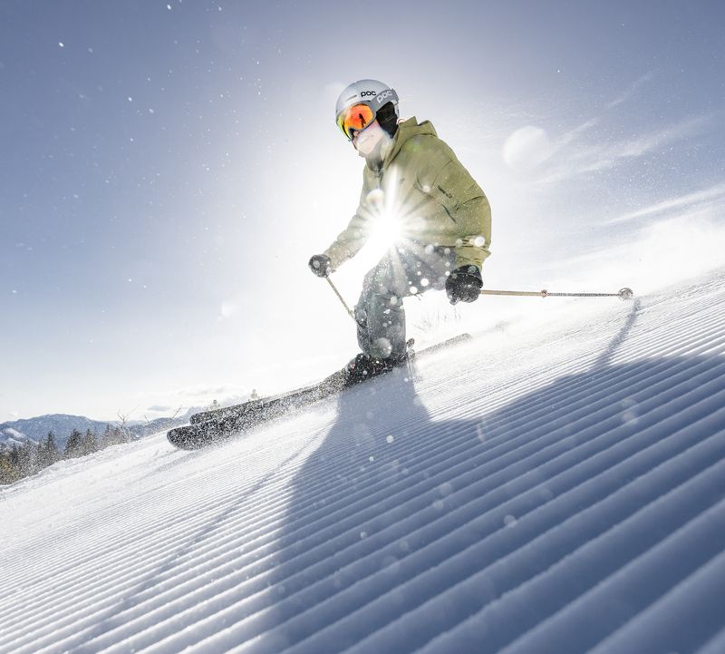 Skier in green jacket skiing dynamically at sunrise over freshly prepared piste, snow crystals flying.