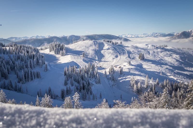 Panoramablick auf eine verschneite Hügellandschaft mit Skipisten, umgeben von Wäldern und Bergen, unter blauem Himmel.