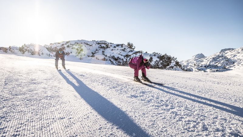 Zwei Skifahrer:innen in Rennhaltung gleiten bei Sonnenaufgang über frisch präparierte Piste am Dachstein Krippenstein
