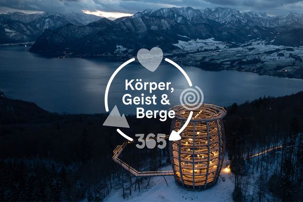 Luftaufnahme eines spiralförmigen Holzturms, der nachts beleuchtet ist, umgeben von verschneiten Bäumen, mit Blick auf einen See und Berge unter einem bewölkten Himmel.