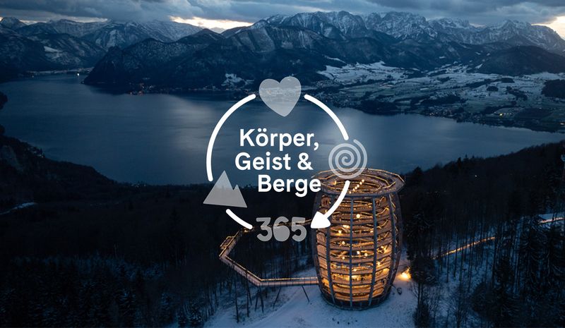 Luftaufnahme eines spiralförmigen Holzturms, der nachts beleuchtet ist, umgeben von verschneiten Bäumen, mit Blick auf einen See und Berge unter einem bewölkten Himmel.