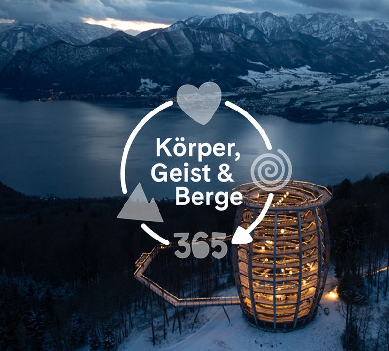 Luftaufnahme eines spiralförmigen Holzturms, der nachts beleuchtet ist, umgeben von verschneiten Bäumen, mit Blick auf einen See und Berge unter einem bewölkten Himmel.