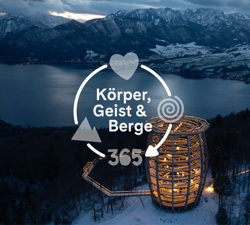 Aerial view of a spiral wooden tower illuminated at night, surrounded by snow-covered trees, overlooking a lake and mountains under a cloudy sky.