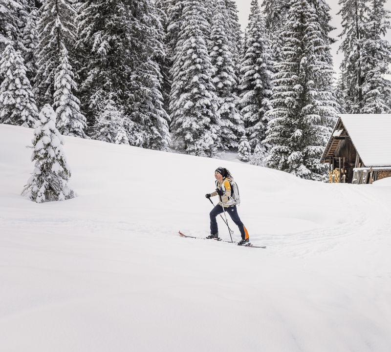 A person cross-country skiing on a snow-covered trail near wooden huts, surrounded by snow-covered trees.